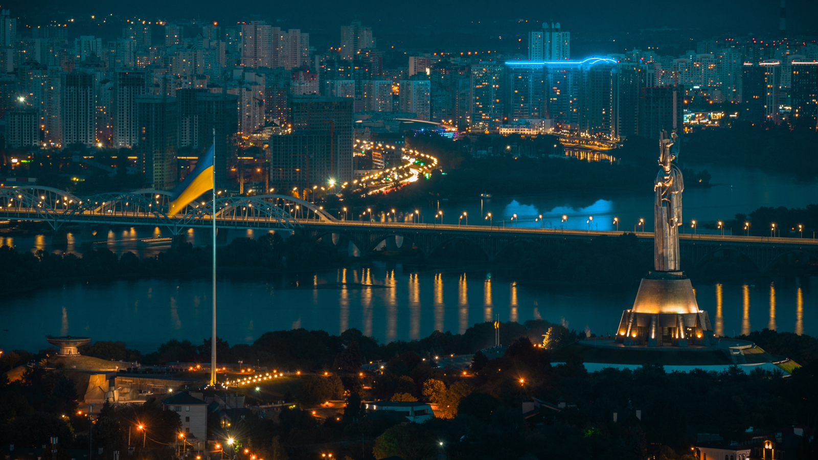 Вид на нічний Київ; окрім іншого, видно прапор України, міст через Дніпро, багатоповерхівки